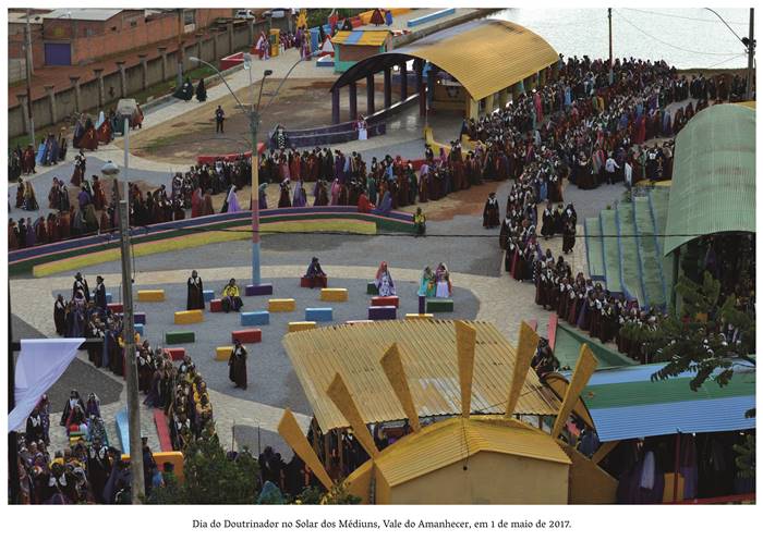  4 m- Dia do Doutrinador no Solar dos Médiuns, no Vale do Amanhecer em 1 de Maio de 2017.. O local é colorido com monumentos triangulados, onde estão reunidos homens e mulheres com vestimentas sacerdotais, em cores diversas. Realização: Academia Planaltinense de Letras, Artes e Ciências (APLAC), pelo Ecomuseu Pedra Fundamental e pelo Coletivo Nativo Audiodescrição produzida  pelo Instituto de Promoção das Pessoas com Deficiência Visual Audiodescritora: Elma Lúcia Rodrigues Consultor: Fernando Rodrigues Este projeto é promovido com recursos do Fundo de Apoio a Cultura do DF